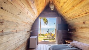 A wooden cabin bedroom with a view of the beach, a bed, a small table, and a heater. - Beachfront Cabins in Bahia – OTX (Porto Seguro)