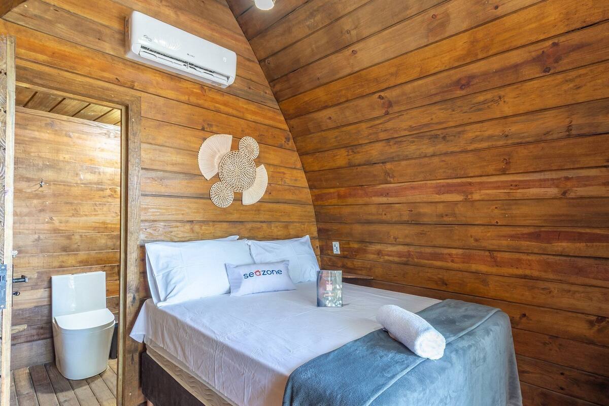 A wooden cabin room with a bed, a small table, a wall-mounted air conditioner, and a wooden door.