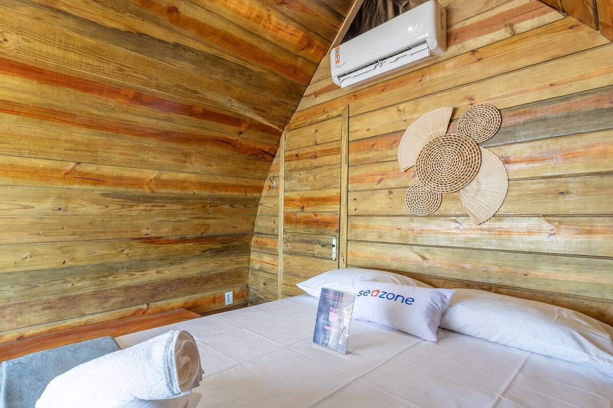 A wooden cabin room with a bed, a wall-mounted air conditioner, and a woven wall decoration.