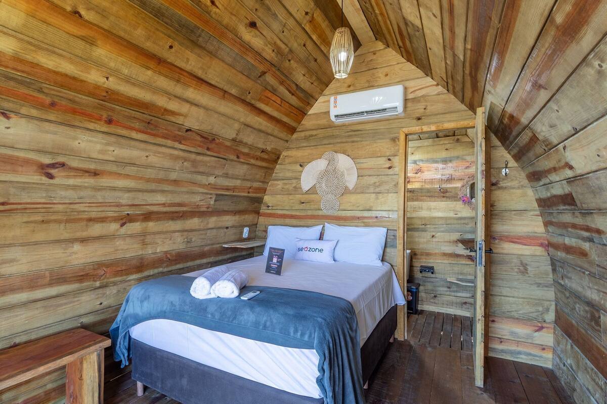 A cozy wooden cabin bedroom with a bed, a wall-mounted air conditioner, a bedside table, and a decorative wall hanging.