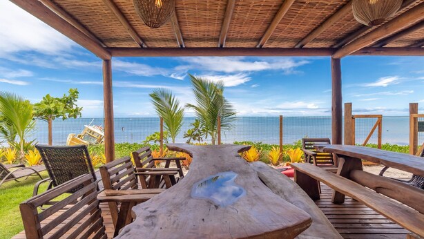 A covered outdoor seating area with wooden benches and tables, overlooking a beach with palm trees and a clear sky. - Beachfront Cabins in Bahia – OTX (Porto Seguro)