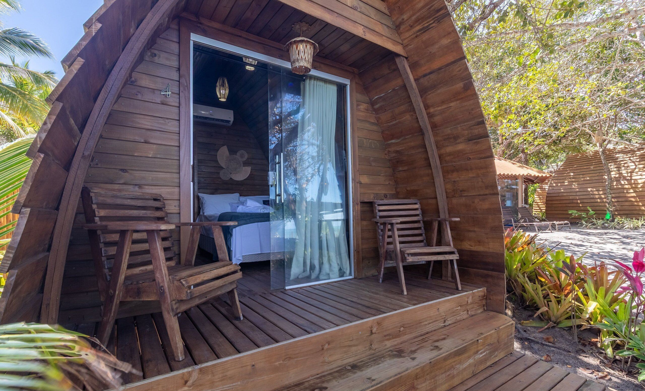 A wooden cabin with a glass door leading to a bed, two wooden chairs, and a small porch.