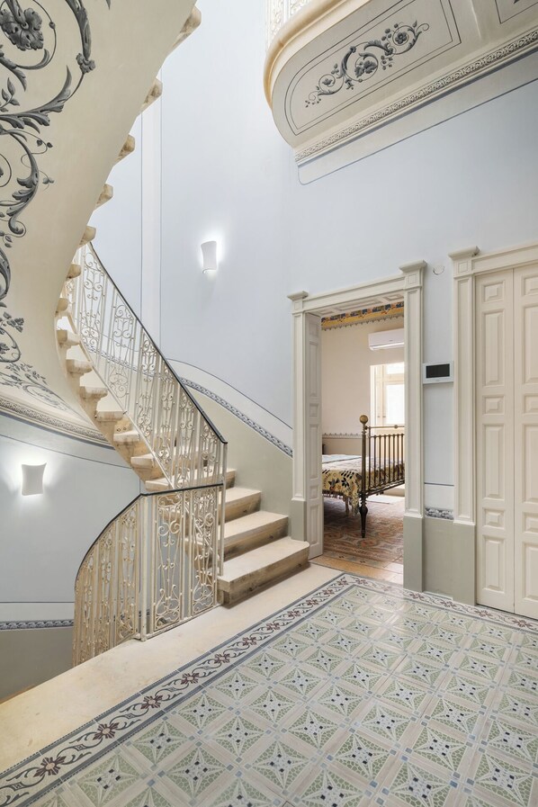 A grand staircase with ornate metal railings and a patterned floor, leading to a room with a bed and a door.