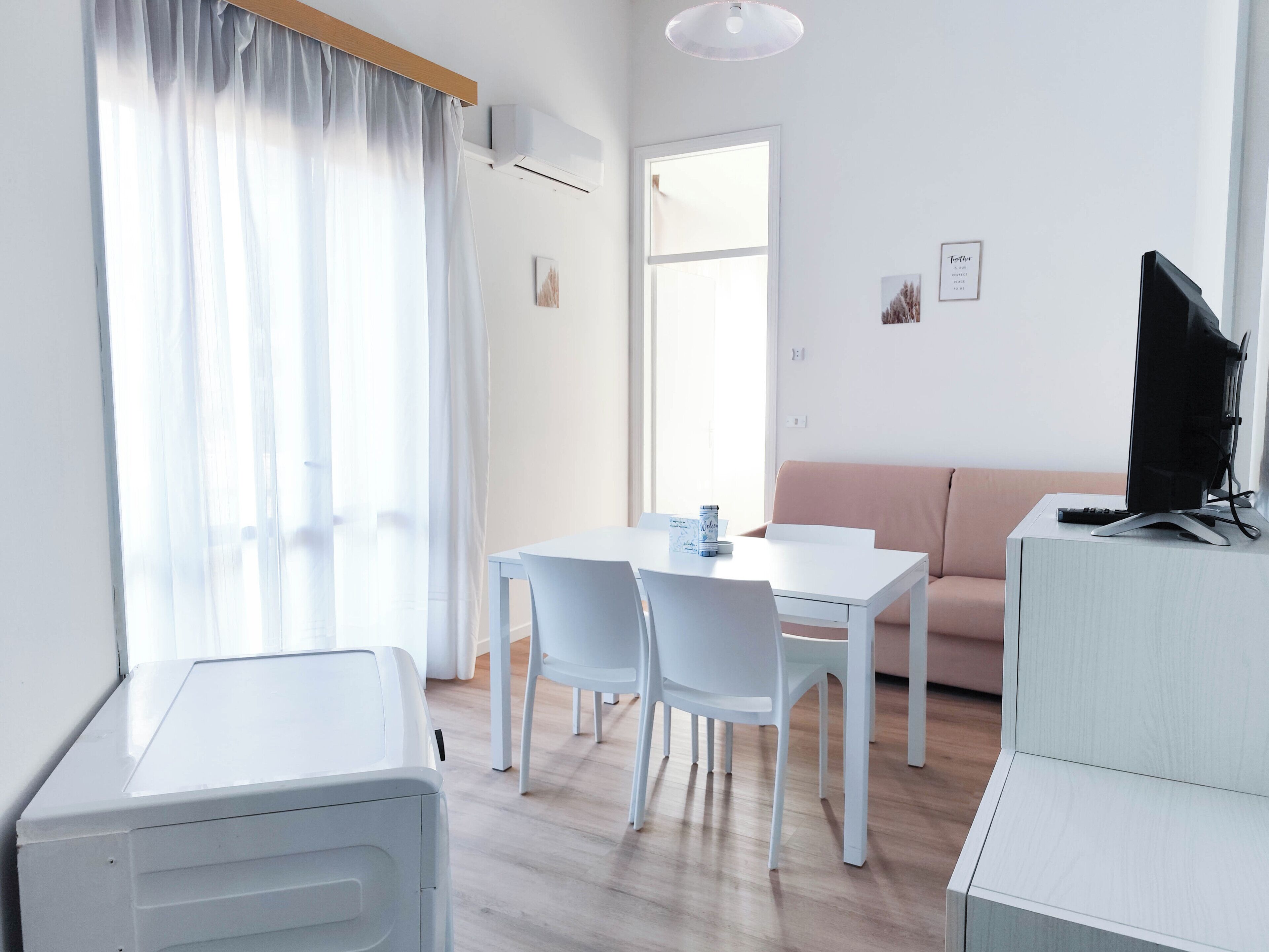Quarto moderno e bem iluminado com mesa de jantar, cadeiras, sofá e TV sobre um móvel.