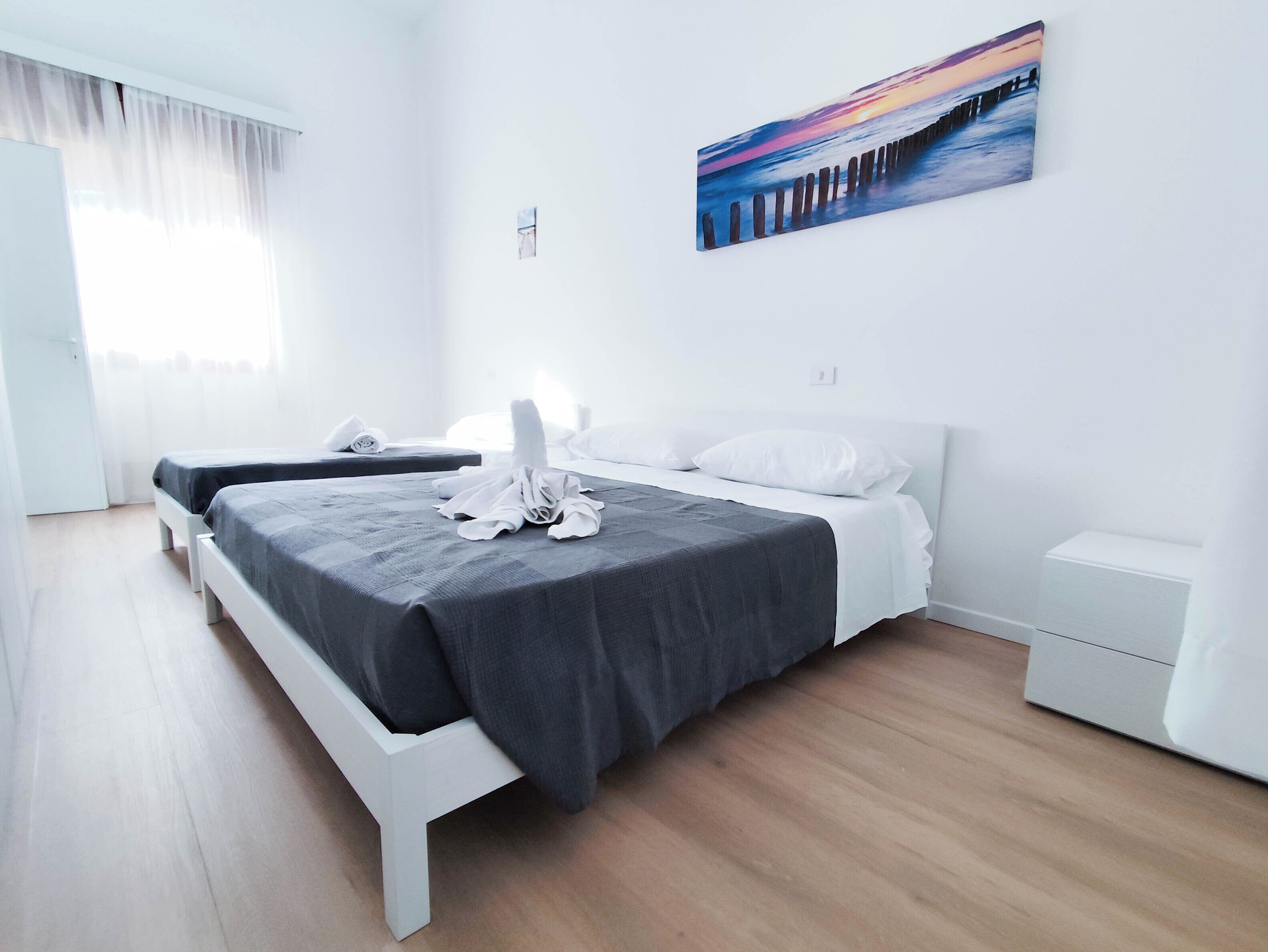 Quarto de hotel moderno com uma cama grande, uma mesa de cabeceira e um quadro com um pôr do sol sobre o oceano.