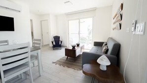 Een moderne woonkamer met een grijze bank, een houten salontafel, een blauwe Adirondack-stoel, en een eethoek met witte stoelen en een houten tafel.