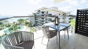 A balcony with a glass table and chairs, overlooking a pool and a building with multiple balconies.