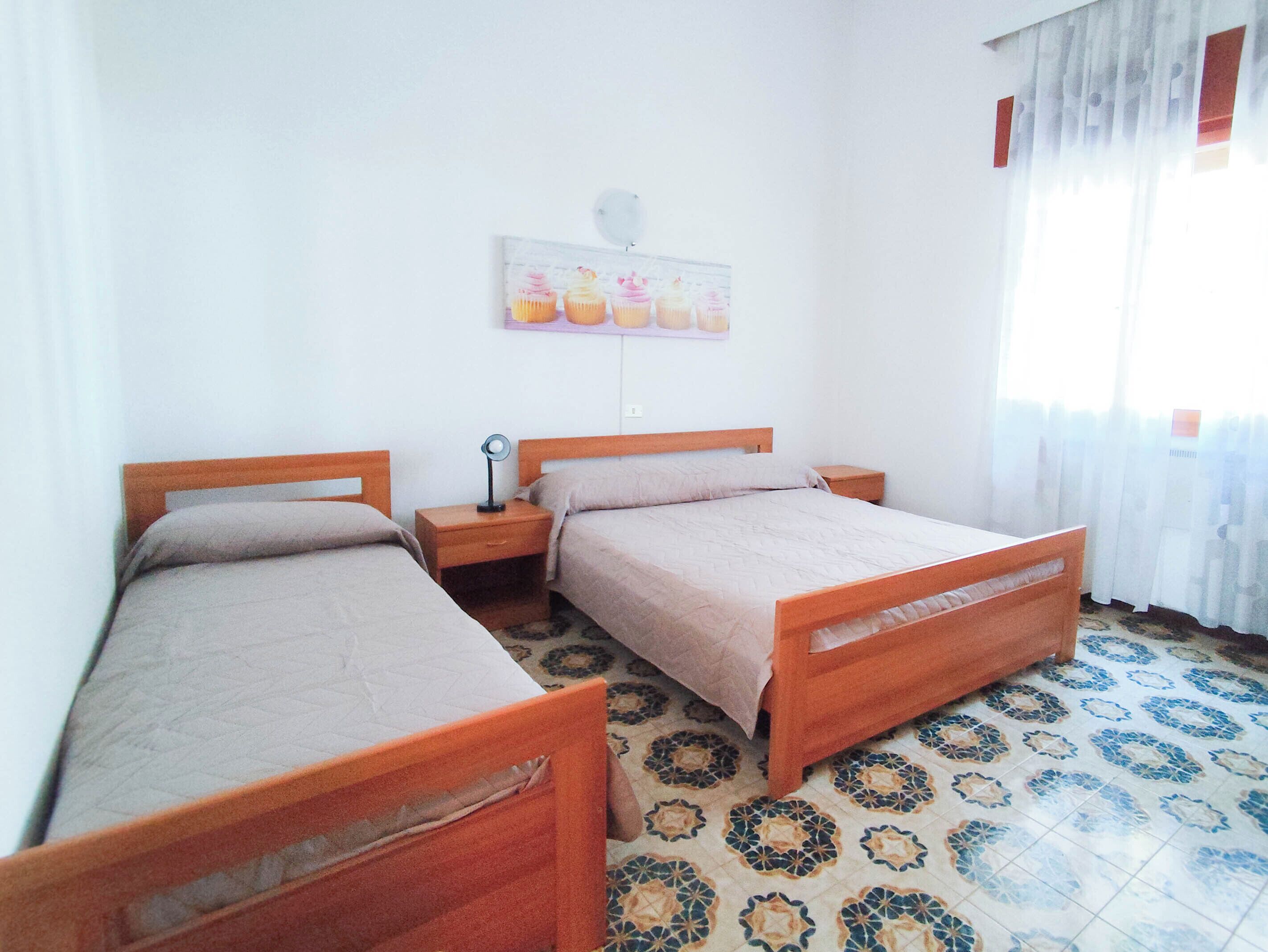 Quarto com duas camas de madeira, uma mesa de cabeceira com abajur, um quadro na parede e ladrilhos no chão com padrão.