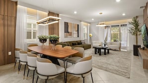 A modern living room with a wooden dining table, white chairs, a sofa, and a coffee table. There are two potted plants, a floor lamp, and a television on a stand. - Mario | Minions | Theater | Private Pool | Disney 15883 (Clermont)