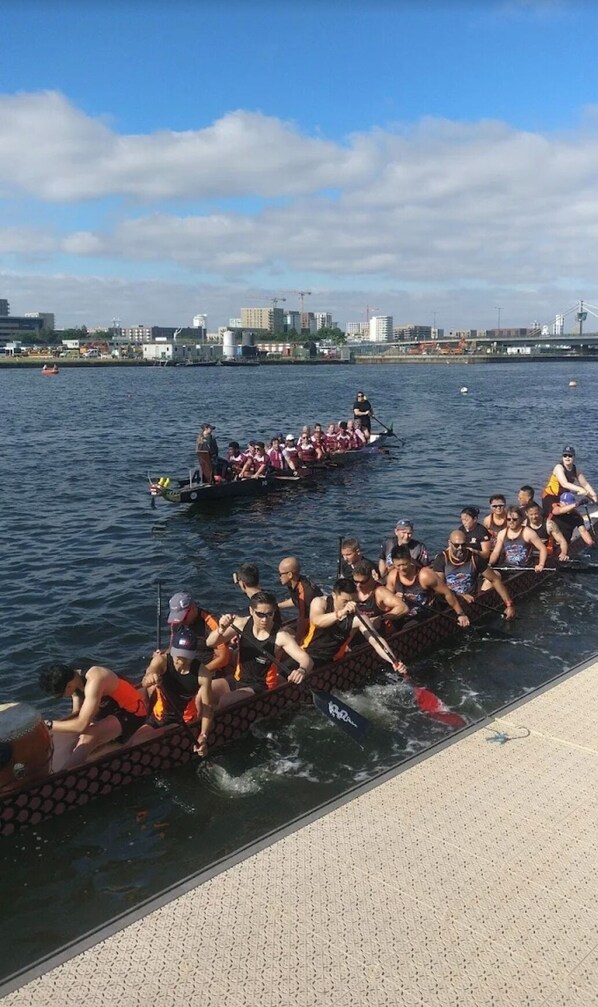 Una carrera de botes dragón con participantes en una embarcación, agua de fondo y un paisaje urbano a lo lejos.