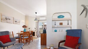A living room with a dining area, a wooden cabinet, and a wall-mounted shelf with decorative items. - Ria's Birds in Cabanas de Tavira – Comfort and Nature by the Ria Formosa (Cabanas de Tavira)
