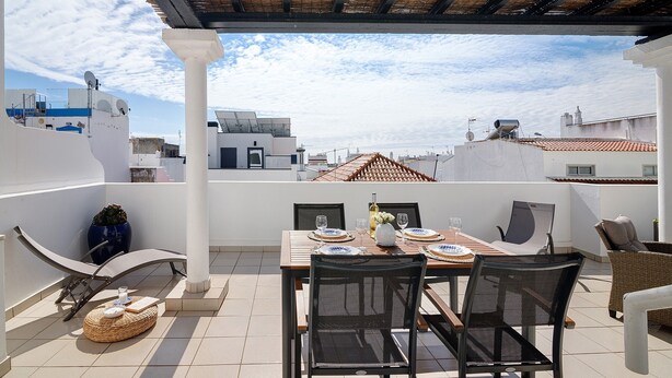 A rooftop terrace with a table set for two, chairs, a potted plant, and a view of the cityscape. - Ria's Birds in Cabanas de Tavira – Comfort and Nature by the Ria Formosa (Cabanas de Tavira)