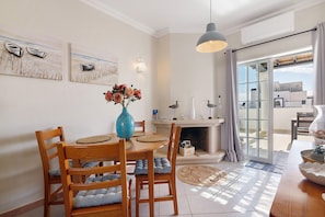 A dining area with a wooden table, chairs, a vase with flowers, and artwork on the wall. - Ria's Birds in Cabanas de Tavira – Comfort and Nature by the Ria Formosa (Cabanas de Tavira)