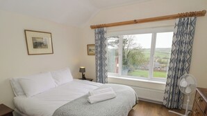 Ein Schlafzimmer mit Bett, Nachttisch, Lampe, einem Fenster mit Vorhängen, Heizkörper, Kommode und Ventilator.