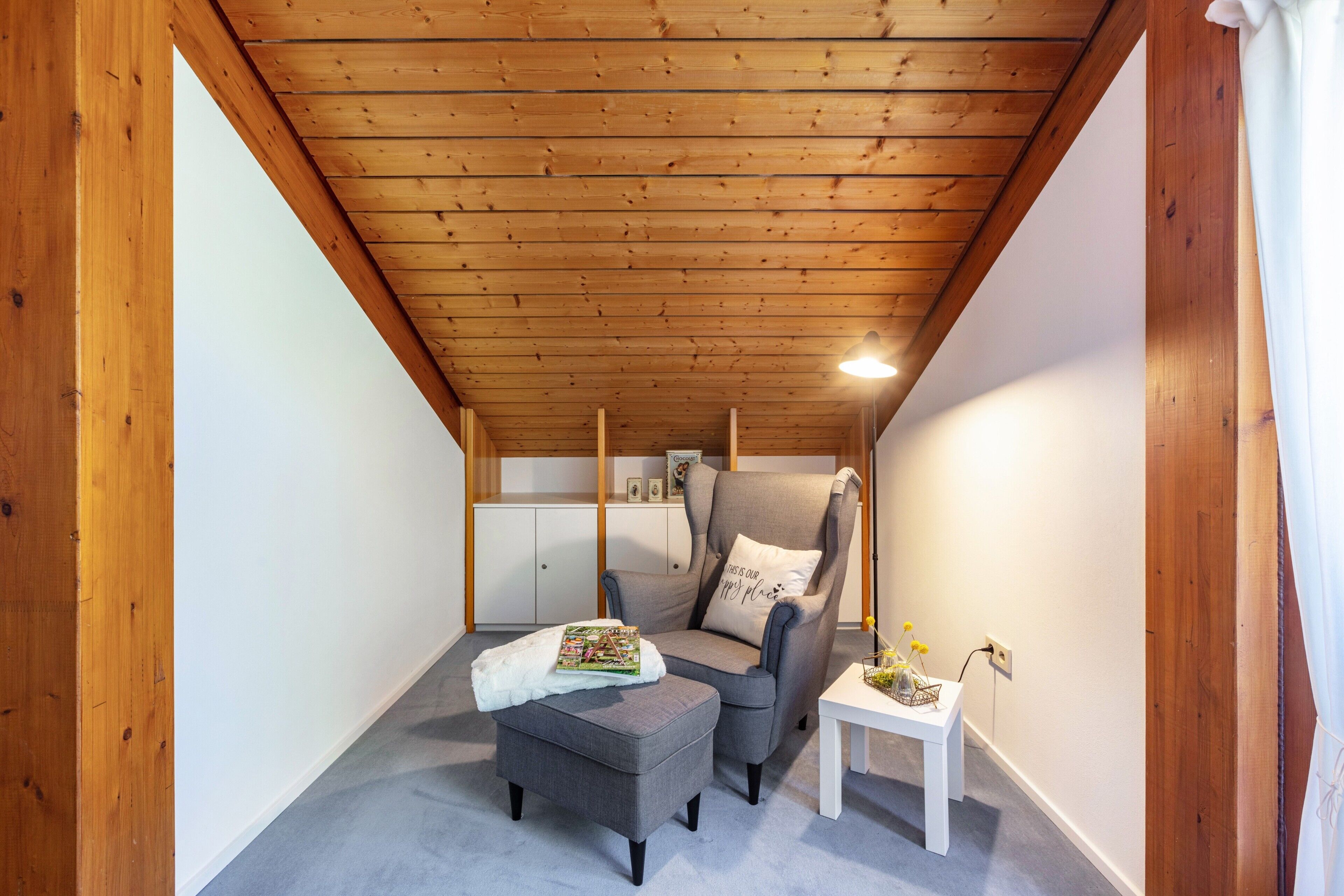 A cozy attic room with a wooden ceiling, a grey armchair, a small ottoman, a white table, and a white cabinet in the background.