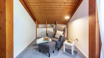 A cozy attic room with a wooden ceiling, a grey armchair, a small ottoman, a white table, and a white cabinet in the background.