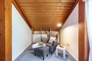 A cozy attic room with a wooden ceiling, a grey armchair, a small ottoman, a white table, and a white cabinet in the background.
