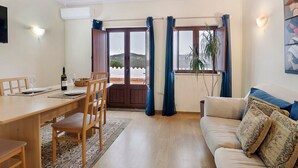 Un salon avec un canapé, une table à manger et des chaises, un téléviseur fixé au mur, et une vue à travers la porte.