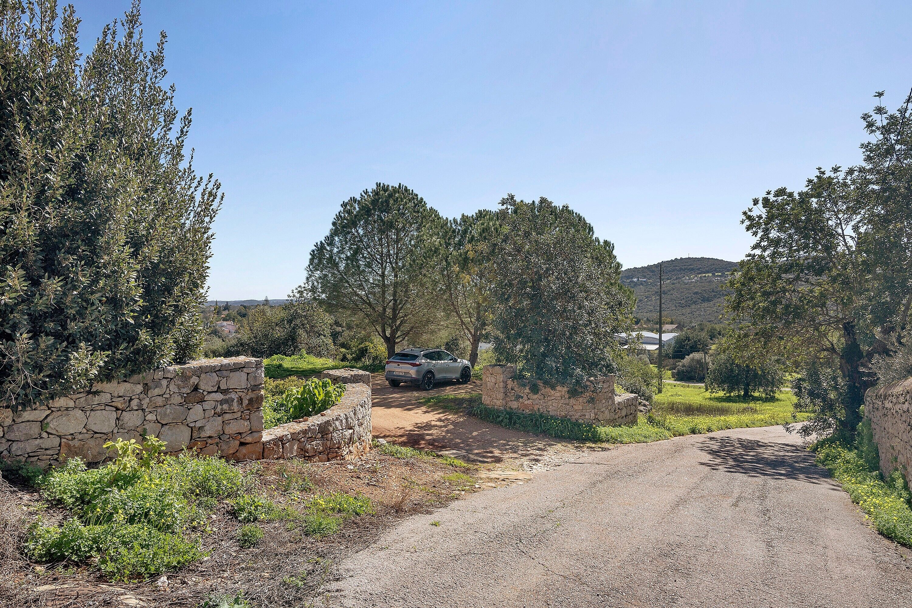 A rural road with a stone wall and greenery on one side, and a car parked on the other.