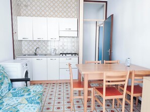 A compact kitchen with white cabinets, a built-in stove, and a sink. There is a dining table with four chairs and a patterned floor. - Cozy Flat Near the Beach (Jesolo)