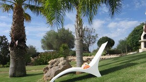 Silla de exterior blanca con cojín, rodeada de césped cuidado, palmeras y un cielo despejado.