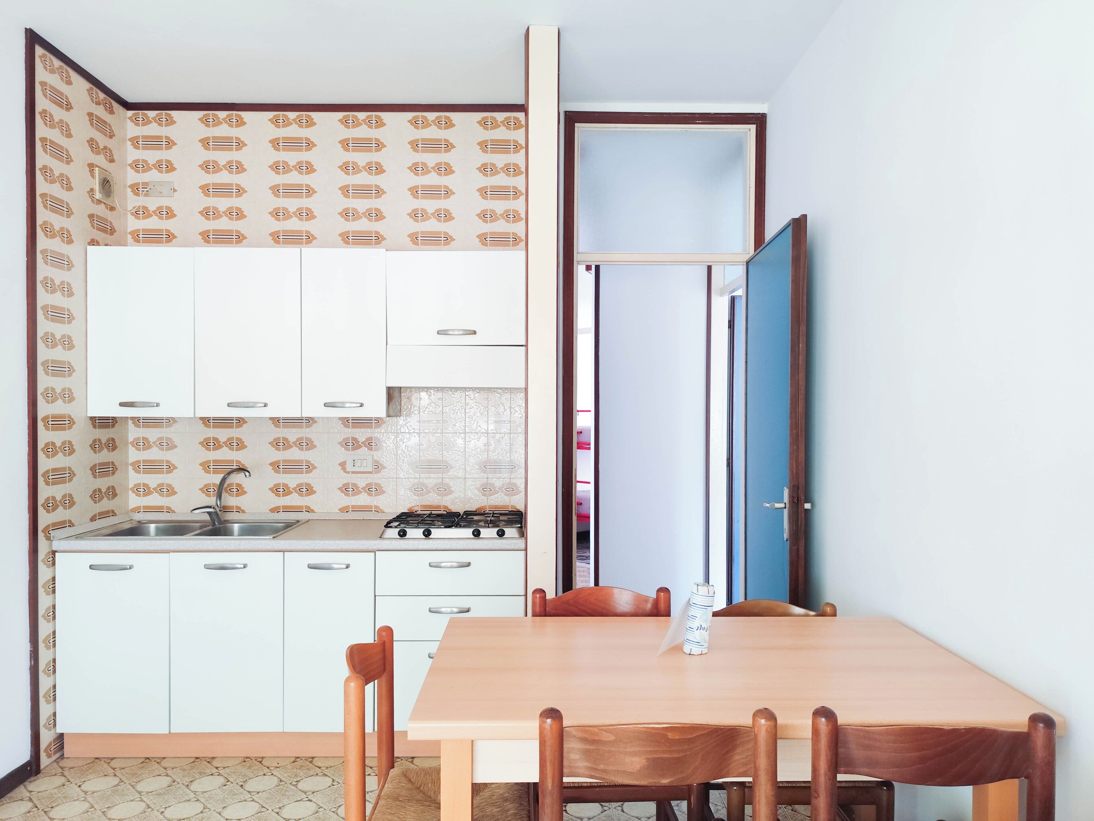 Cozinha compacta com armários brancos, fogão a gás e pia. Há uma mesa de jantar com cadeiras e uma porta azul ao fundo.