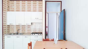 Cozinha compacta com armários brancos, fogão a gás e pia. Há uma mesa de jantar com cadeiras e uma porta azul ao fundo.