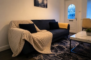 A cozy living room with a dark sofa, a cream-colored throw blanket, and a small table with a plant.