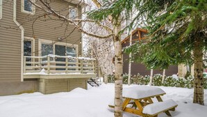 A snow-covered outdoor area with a wooden bench and a picnic table, a wooden deck, and a building with windows and a clock. - Hôtel à la maison - Chez Charcole (Saint-Ferréol-les-Neiges)