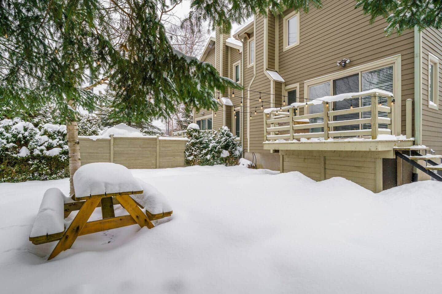 A snow-covered deck with a wooden chair, a wooden fence, and a building with windows and a balcony.