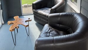 Ett rum med två fåtöljer i läder, ett soffbord i trä och en utsikt genom ett välvt fönster mot ett hus och en gata.