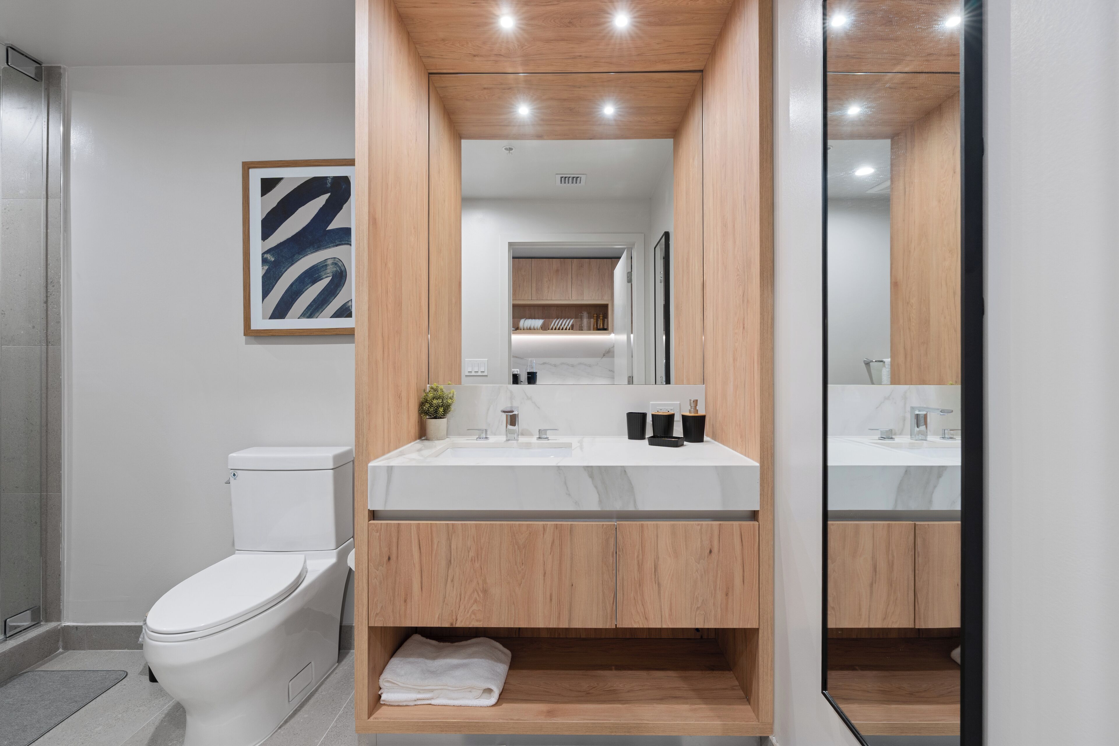 A modern bathroom with a white toilet, a wooden vanity with a mirror, and a framed abstract artwork on the wall.