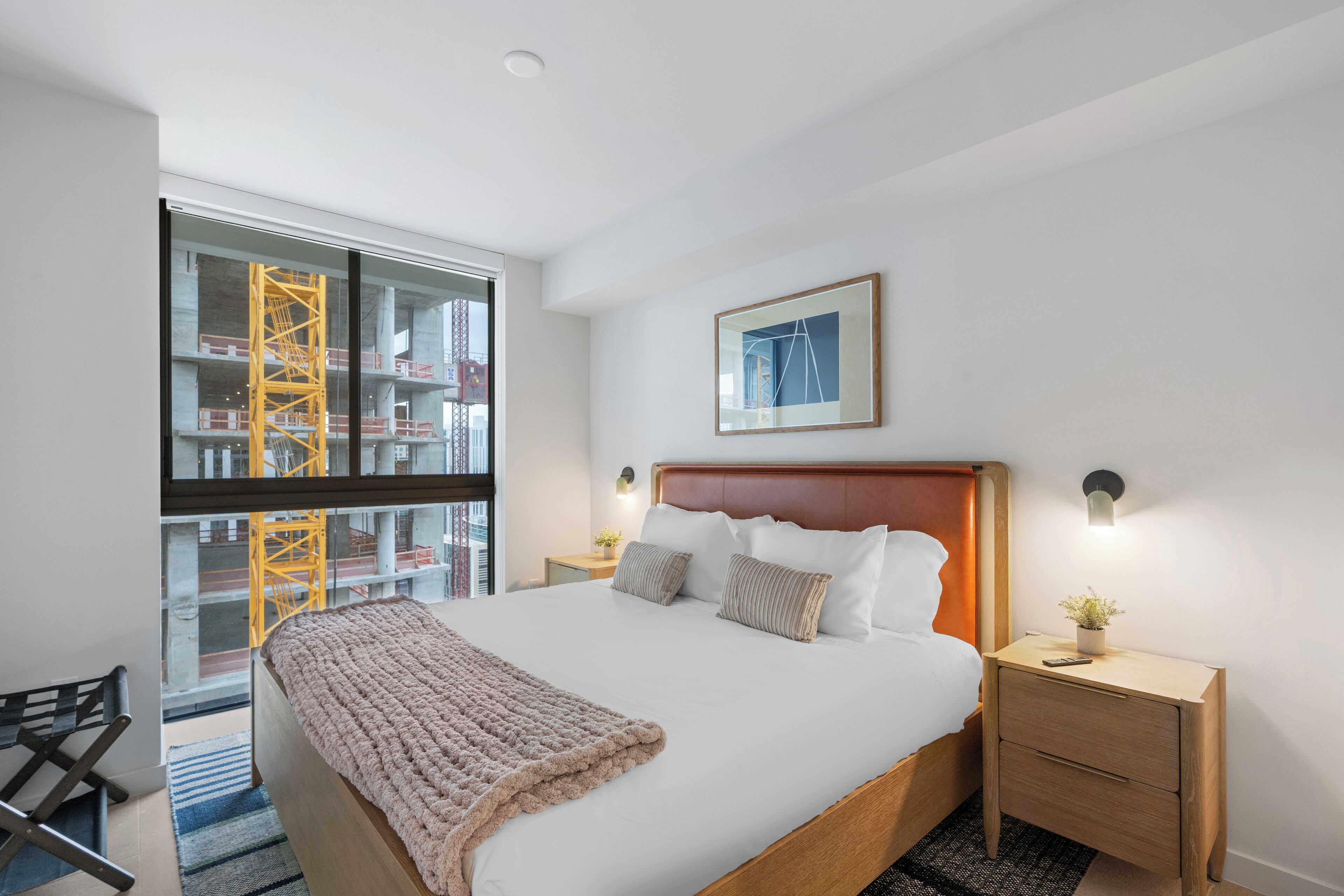 A modern hotel room with a large bed, a wooden nightstand, a framed picture on the wall, and a view of a construction site through a glass door.