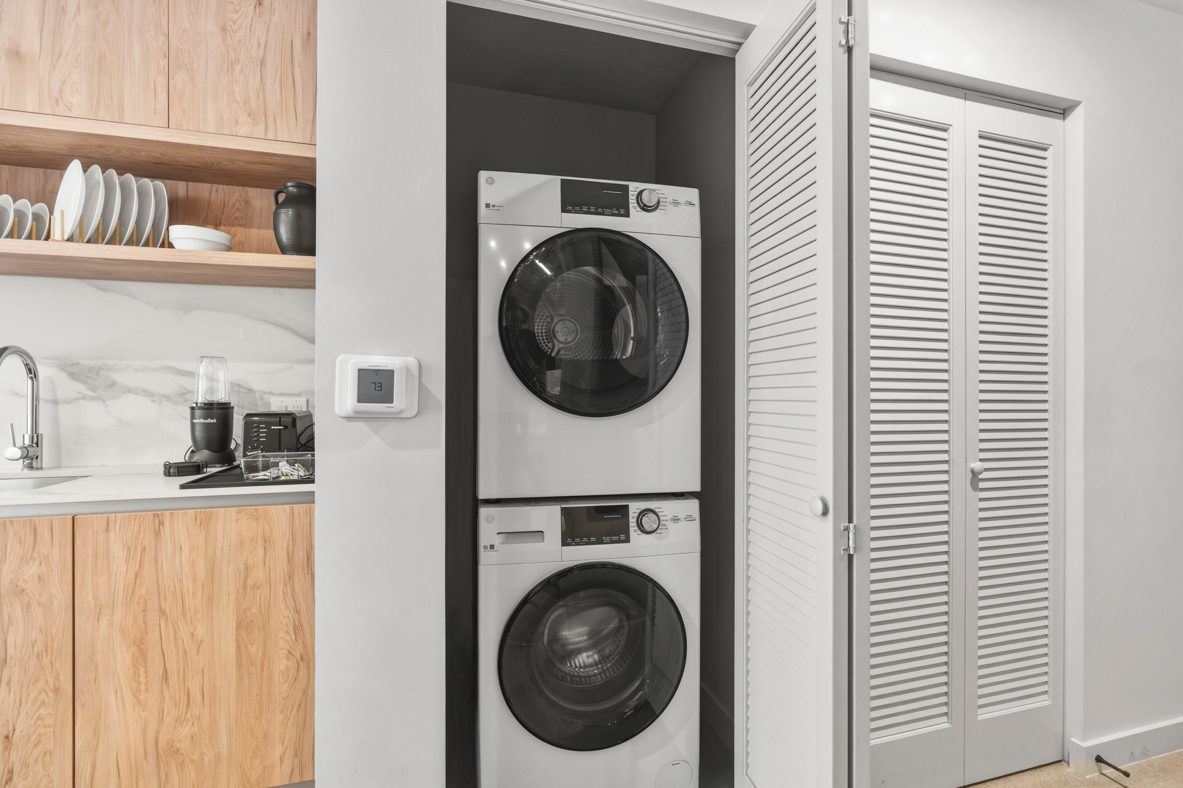 A laundry room with stacked washers and dryers, a coffee maker, and a shelf with dishes.