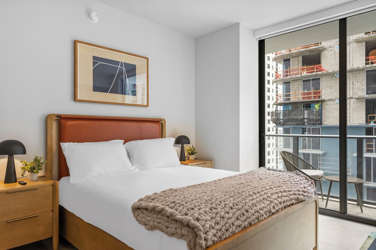 Une chambre d’hôtel moderne avec un grand lit, une table de chevet avec une lampe, un tableau encadré au mur et une vue sur un bâtiment en construction.