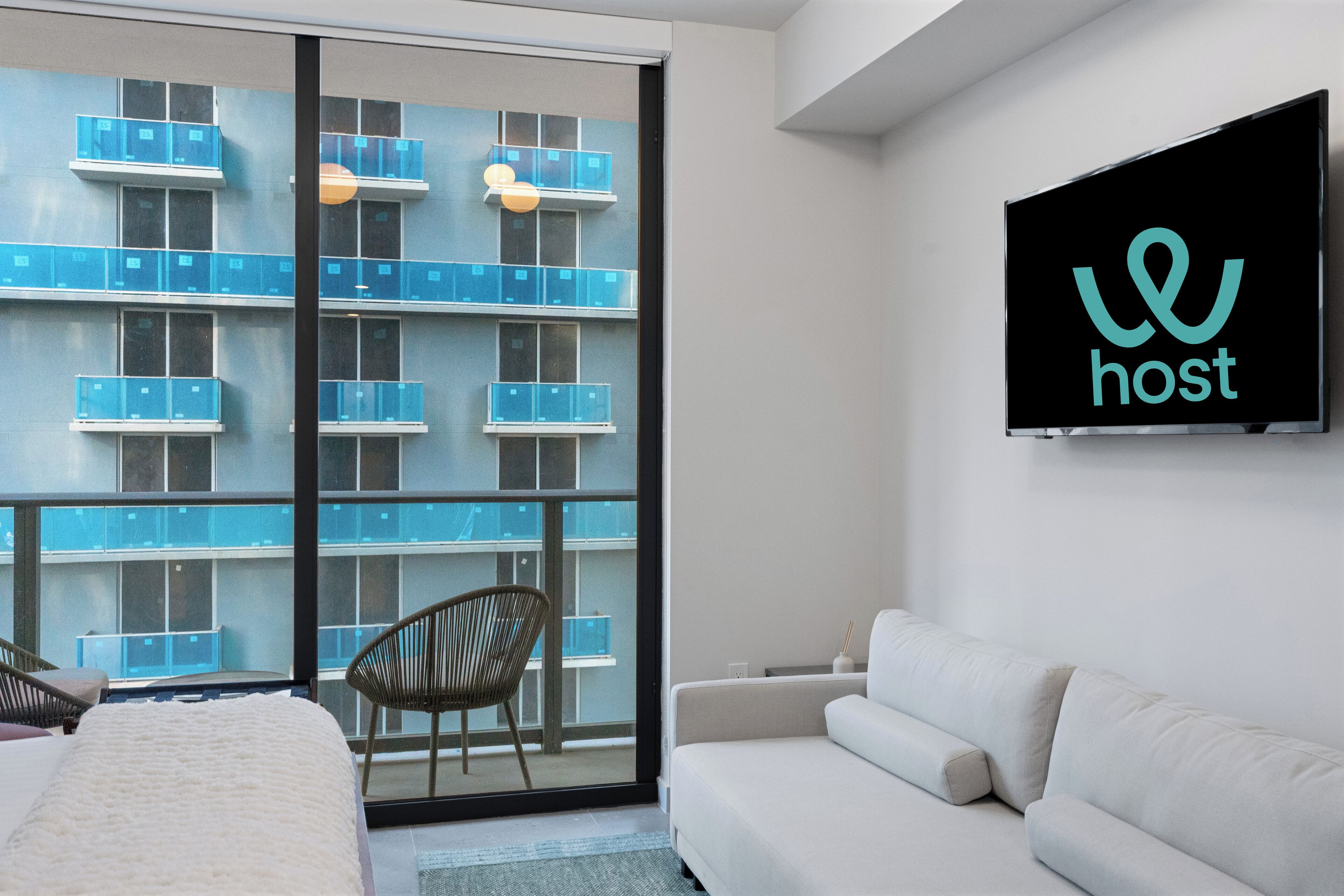 A modern hotel room with a large sliding glass door leading to a balcony, a white sofa, a bed with a white cover, and a television mounted on the wall displaying the word "host".