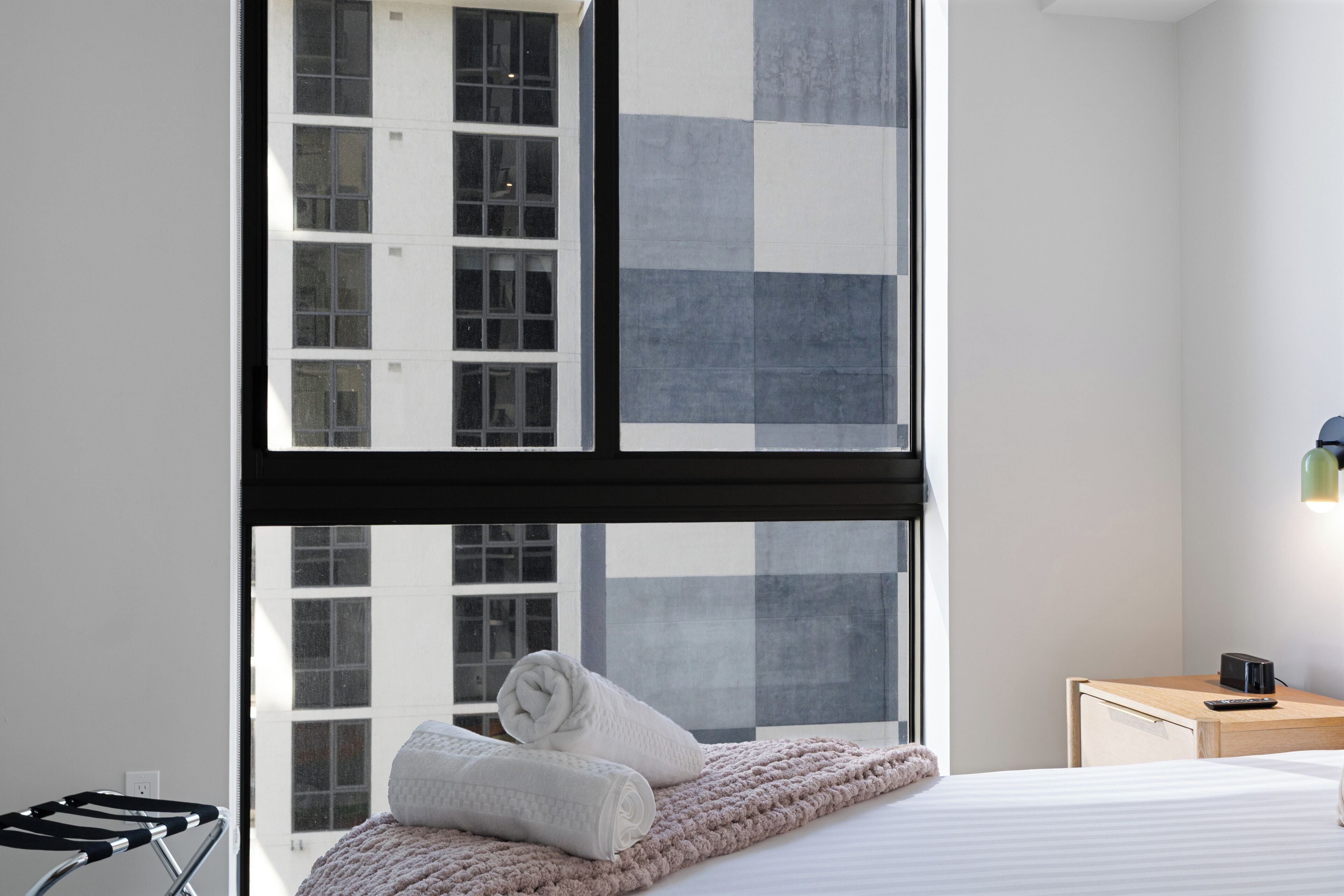 A modern hotel room with a large window, a bed with a white comforter, a bedside table with a lamp, and a view of a building with multiple windows.