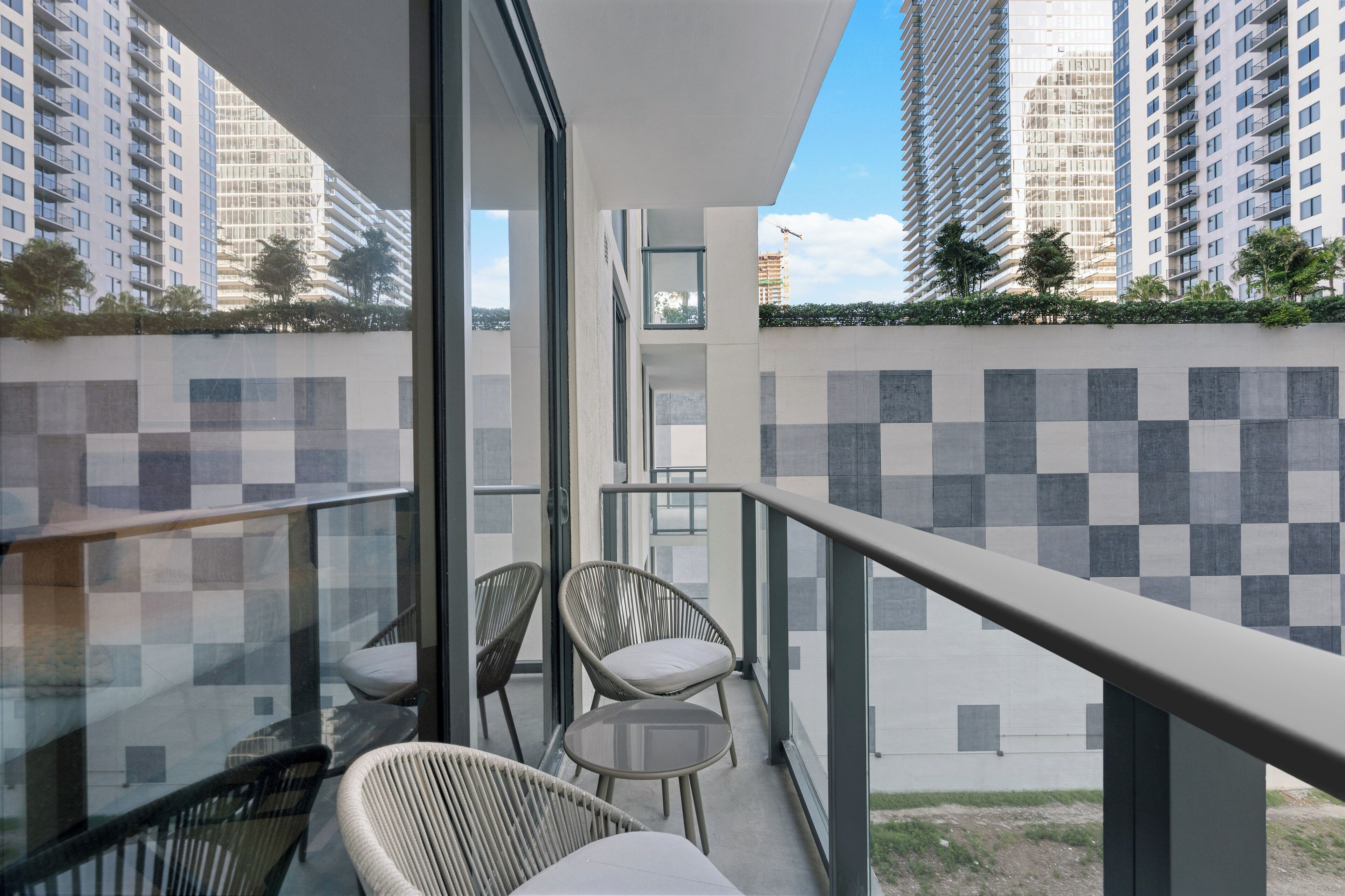 A balcony with two wicker chairs and a small table, overlooking a cityscape with tall buildings and a construction crane.