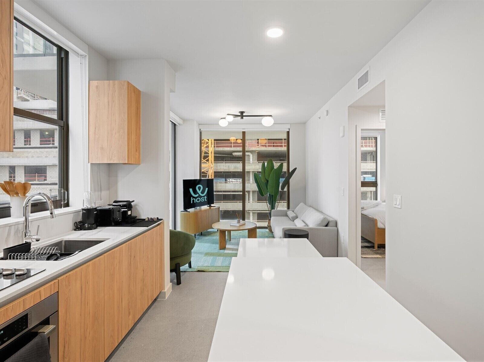 A modern kitchen with a built-in oven, a sink, and a coffee machine. There is a dining area with a white table and a sofa.