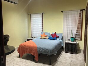 Quarto com cama, mesas de cabeceira, uma cadeira, planta em vaso e janela com cortinas.