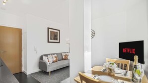 Ein modernes Wohnzimmer mit einem grauen Sofa, einem gerahmten Bild an der Wand und einem Essbereich mit Tisch für zwei Personen.