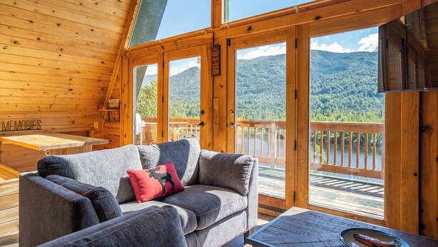 A wooden cabin interior with a sofa, a table, and a view of a lake and mountains.