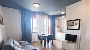 A modern living room with a sofa, dining table, chairs, kitchenette, and a TV. - Cozy Studio Near the Beach (Jesolo)