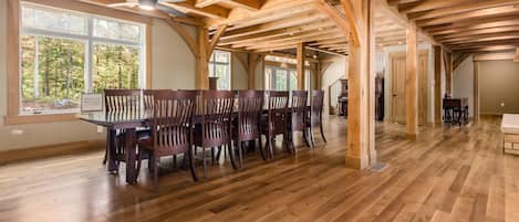 A spacious dining area with a long wooden table and chairs, large windows with a view of trees, and a high ceiling with exposed wooden beams.