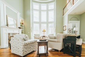 A living room with floral-patterned sofas, a fireplace, a large bay window, and a staircase. - Hocking Manor | Hocking Hills, 8BR, Indoor Pool (Rockbridge)
