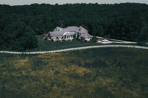 A large house surrounded by a white fence, set on a grassy hill with dense forest in the background. - Hocking Manor | Hocking Hills, 8BR, Indoor Pool (Rockbridge)