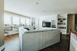 A modern living room with a grey sofa, white built-in shelving, a flat-screen TV, and a staircase with a wooden railing. - Hocking Manor | Hocking Hills, 8BR, Indoor Pool (Rockbridge)