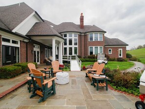 A covered patio area with Adirondack chairs, a barbecue grill, and a stone-tiled floor. - Hocking Manor | Hocking Hills, 8BR, Indoor Pool (Rockbridge)
