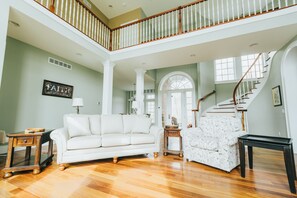 A spacious living room with a white sofa, a floral armchair, and a black coffee table. A wooden staircase with a white railing leads to an upper level. - Hocking Manor | Hocking Hills, 8BR, Indoor Pool (Rockbridge)