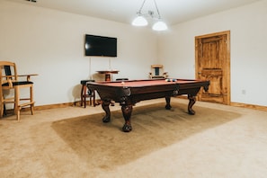 Une pièce comprenant une table de billard, deux chaises, un téléviseur fixé au mur et une porte en bois.